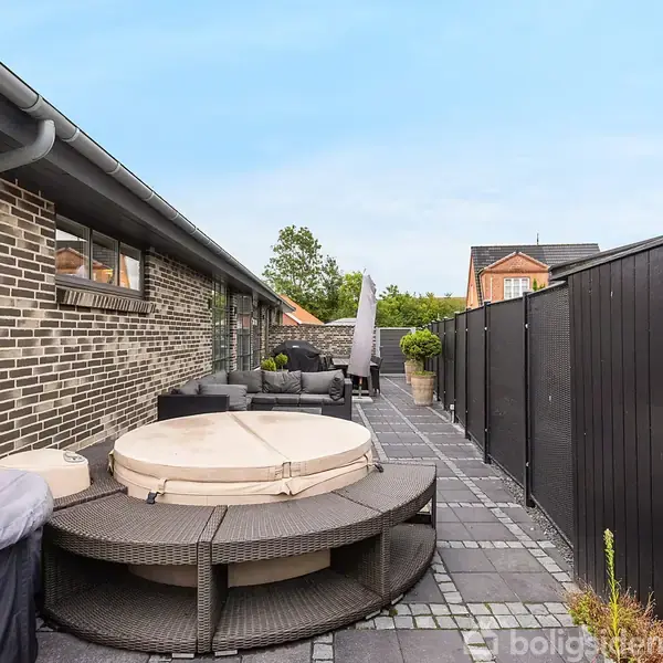 Et rundt vildmarksbad står stille på en terrasse med grå fliser. Til venstre er en murstensvæg, og til højre en sort hegn. I baggrunden er der udendørs møbler.