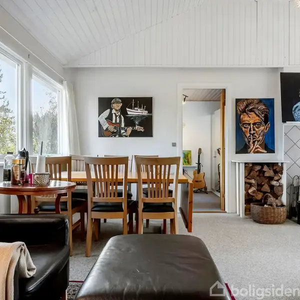 Stue med sort lænestol og lille bord, spisebord med seks stole. Væggene prydet med tre malerier. Pejs op mod væggen med brændestabel ved siden af. Store vinduer til venstre.