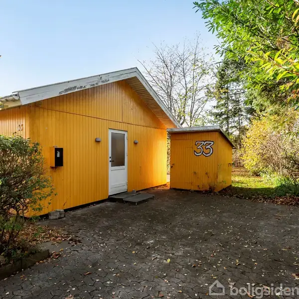 Gul træbygning med hvid dør står stille på en brolagt gårdsplads omgivet af buske og træer. Et mindre skur til højre viser tallet "33".