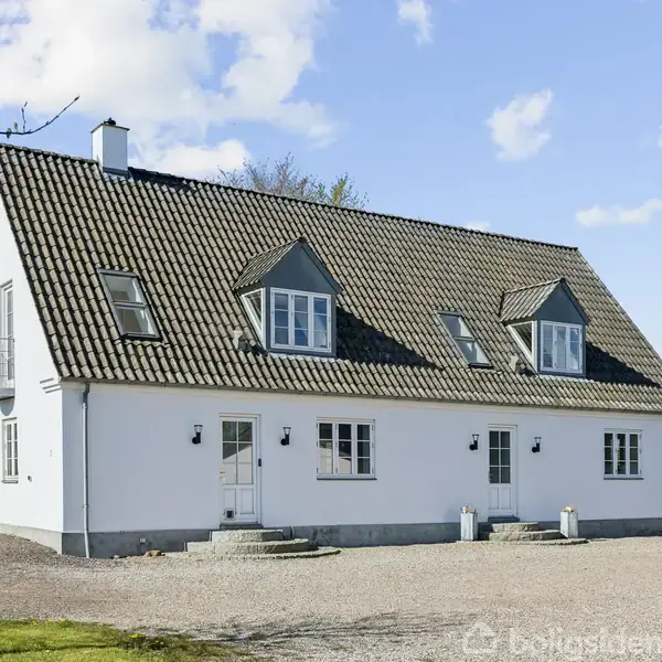 Hvidt, to-etagers hus med sorte skråtag og kvistvinduer står på en grusbelagt grund. Rundt omkring er en grøn plæne og sparsomme træer under en klar blå himmel med skyer.
