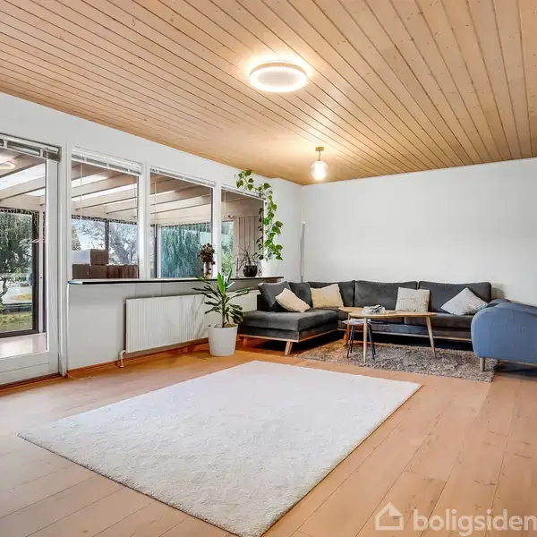 Stue med hjørnesofa ved siden af en brændeovn. Store vinduer til venstre viser en terrasse og have. Lys trægulv og -loft med planter og tæppe som udsmykning.