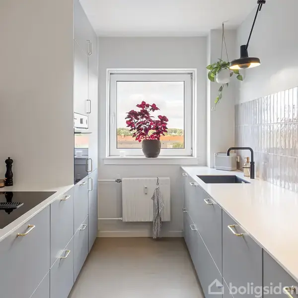 Et moderne køkken med grå skabe på hver side. En plante står i vinduet. En håndvask er til højre, loftslamper hænger over bordet, og væggene har flisebelægning.