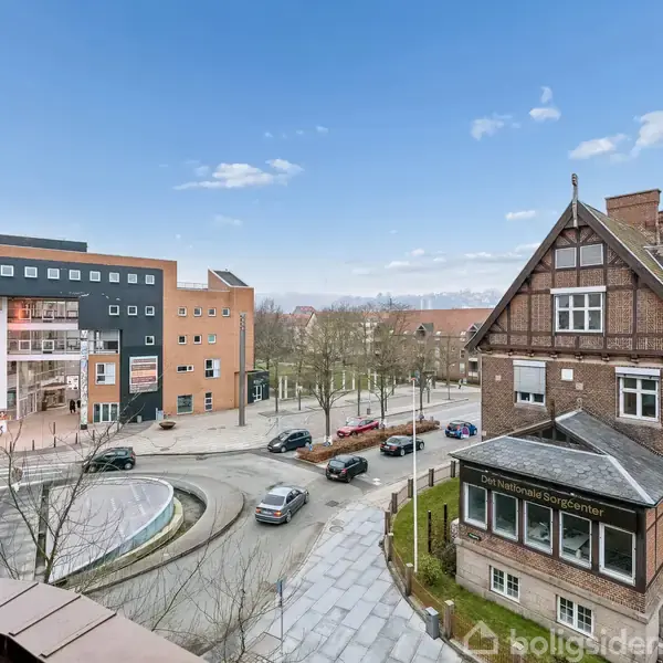 Historisk murstensbygning står ved siden af en rund vej med biler. I baggrunden ses et moderne kulturhus med tekst "VIBY BIB" på siden. Det er en bymiljøscene.