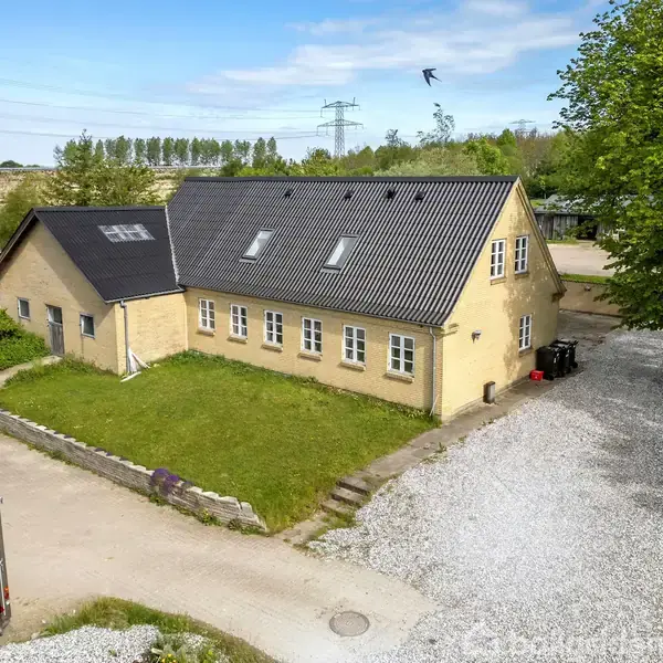 Et gult hus med sort tag står omgivet af en grusvej og træer. En fugl svæver i luften, og der er åbne marker og elektriske master i baggrunden.