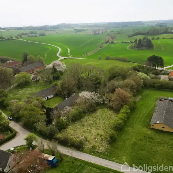 Landsbyområde med spredte huse omkranset af skove og marker; snoede veje fører gennem den frodige grønne landskab, med en vindmølle synlig i baggrunden på en overskyet dag.
