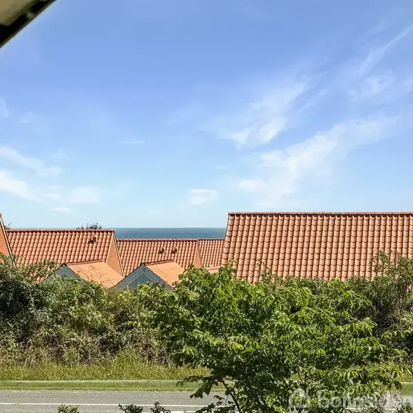 Røde tegltage ligger stille mod en blå himmel, med grøn vegetation i forgrunden og havet synligt i horisonten.
