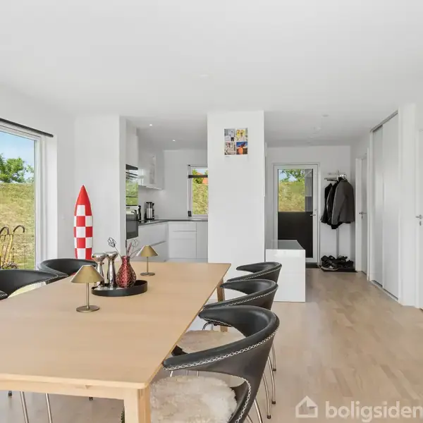 Spisebord med sorte stole er centreret i et moderne, lyst rum. På bordet står pynt og lamper. Bagved ses en køkkenafdeling med vinduer, der viser en grøn græsplæne udenfor.