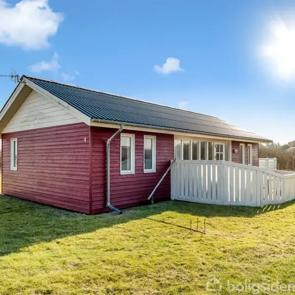 Et rødt træhus med sort tag står på en græsplæne. Huset har vinduer og en hvid veranda. Solen skinner, og himlen er blå med spredte skyer.