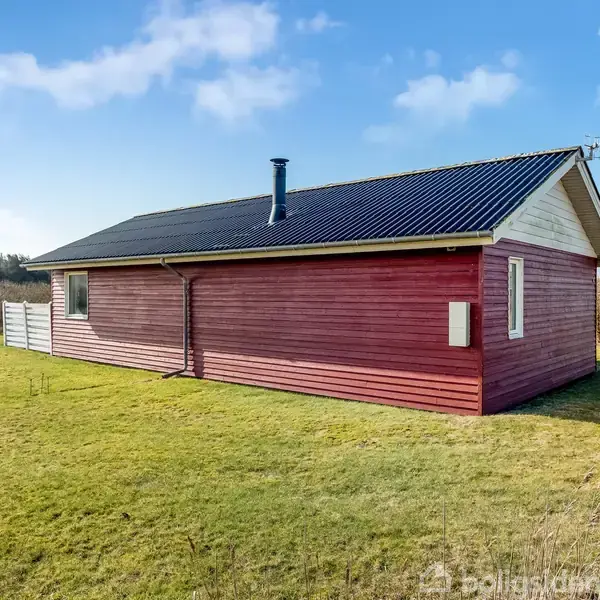 Et rødt træhus med sort tag står på en grøn græsplæne. Skorsten rager op fra taget. I baggrunden ses en hæk og blå himmel.