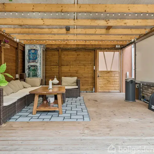 Siddeområde med hjørnesofa og træbord, omgivet af overdækket terrasse med trægulve og indrammet af trævægge. En projektorskærm hænger til højre, dekoreret med planter og kunst.