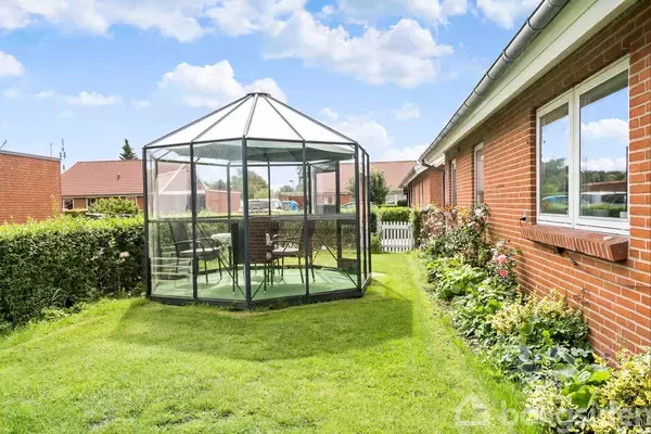 Drivhus med glasvægge indeholder to stole og et bord, placeret på en græsplæne ved siden af en rød murstensbygning med vinduer. Solrigt vejr og blå himmel danner baggrunden.