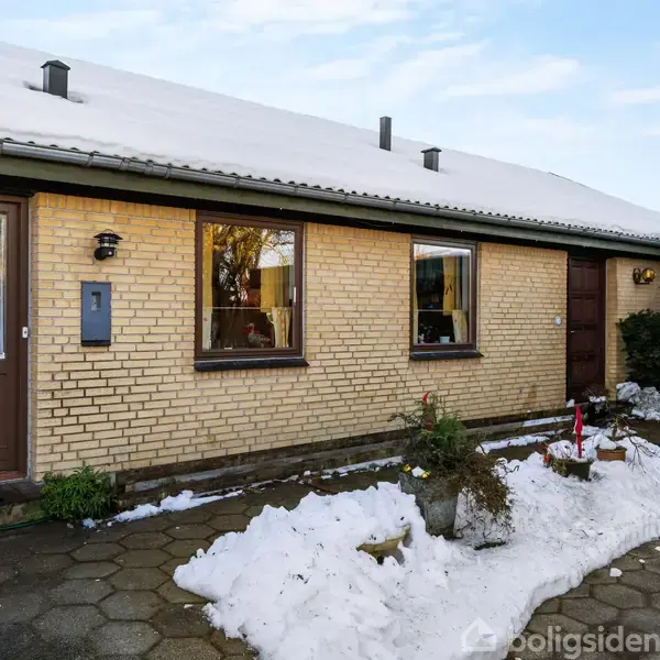 En gulstensbygning med sne på taget står stille, omgivet af spredt sne i forhaven. Der ses en grå indkørsel og et lyst overdækket område til højre.