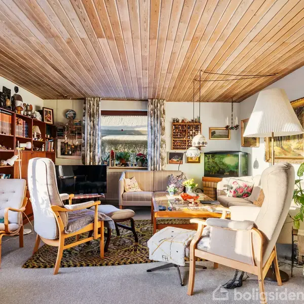 Stue med polstrede træstole omkring et sofabord, en reol med bøger og dekorationer i baggrunden, og store vinduer med gardiner. Indretningen er lys og hjemlig med planter.