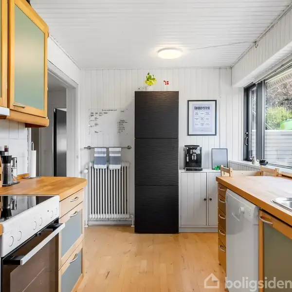 Køkken med træbordplader, hvide skabe og moderne kogeplade; høj sort køleskab ved siden af en kaffemaskine. Store vinduer giver udsigt til have. Tekst på opslagstavle: "INDKØBSLISTE".