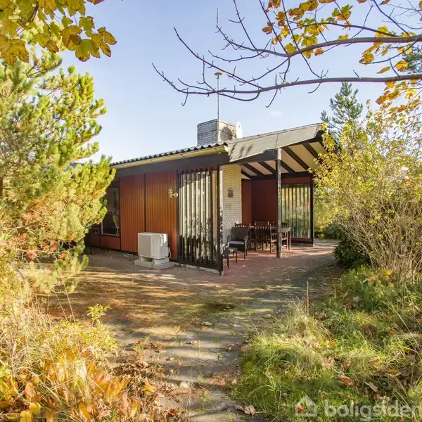 Et sommerhus står alene i en frodig have, omgivet af træer og buske. Huset har træbeklædte vægge og en udendørs terrasse med borde og stole.