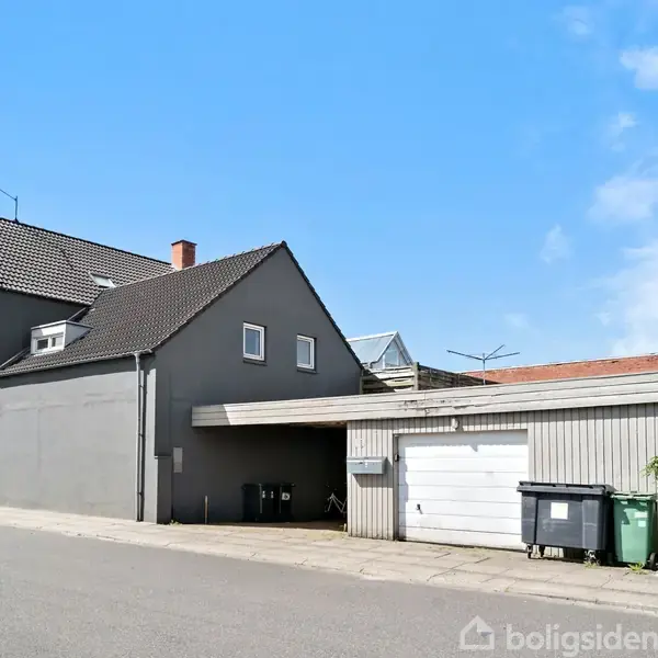 Et gråmalet hus med skråt tag står ved en vej. Tilknyttet er en garage med en hvid port, omgivet af skraldespande. Vejret er solrigt med blå himmel.