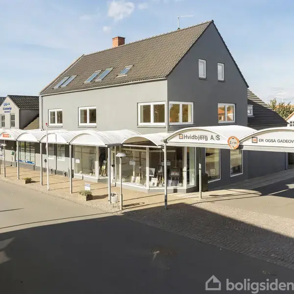 En grå, to-etagers bygning med store vinduer under en række hvide markiser. Skilte: "Elindbjerg A/S" og "Er ogsa Gasdverejforening". Omgivet af en stille gade og fortov.