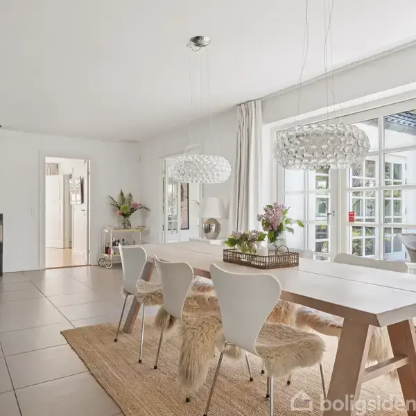Spisebord med hvide stole og pelsvattetæpper står i en lys spisestue med store vinduer. En moderne brændeovn og indbydende indretning skaber en hyggelig atmosfære.