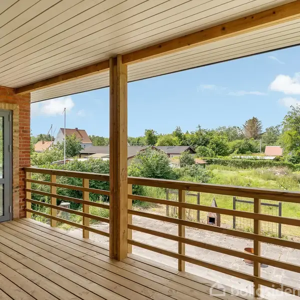 En overdækket terrasse med trægulv og rækværk, der åbner sig mod en frodig have med grønne træer og huse i baggrunden under en blå himmel med få skyer.