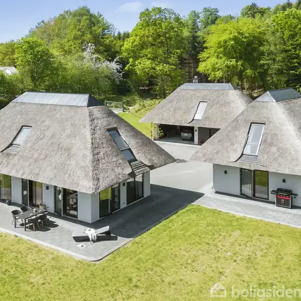 Tre huse med stråtag står i nærheden af hinanden på en velplejet græsplæne omgivet af frodig skov. Møbler og en jacuzzi findes udenfor på en terrasse.