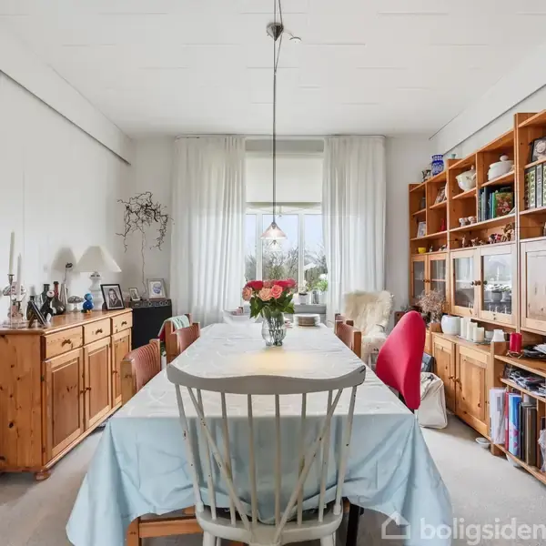 Et spisebord dækket med en lys dug står centralt med blomster i en vase. Omkring er der træskænke og bogreoler fyldt med bøger, lys og dekorationer i en lys spisestue.