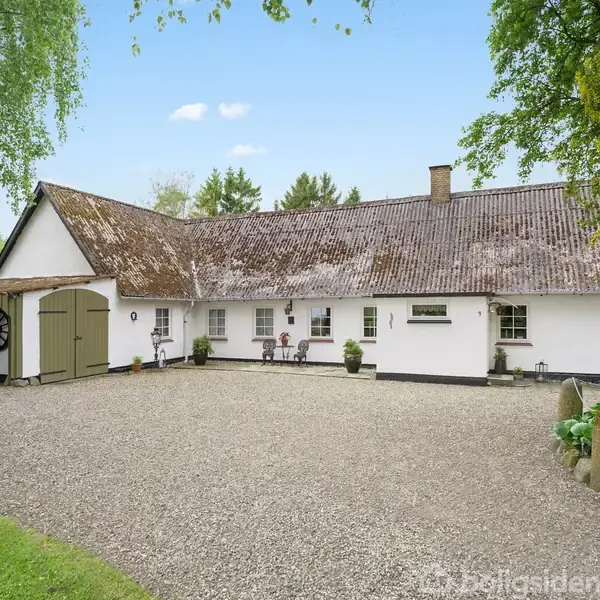 Et landligt hus med hvid facade og skråt tag står i en grusbelagt gårdsplads omgivet af grønne træer. Der er en garage med en bil til venstre.