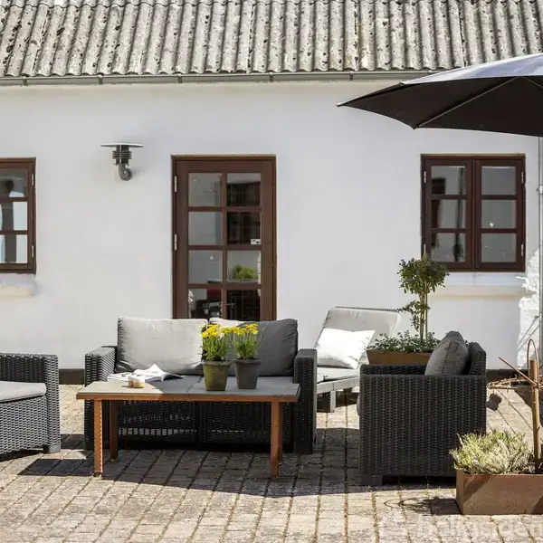 Udendørs terrassemøbler står på en brolagt gårdsplads foran en hvid bygning med brune vindueskarme. En sort parasol skaber skygge. Blomsterpotter med planter tilføjer farve.