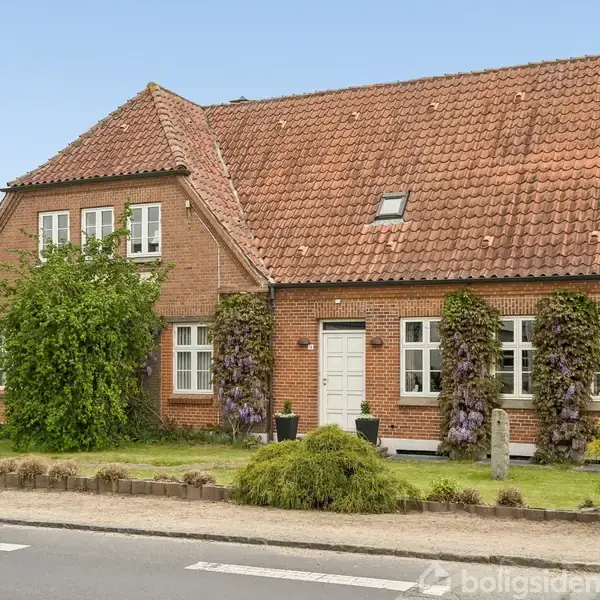 Et rødt murstenshus med stejlt, teglbelagt tag og klatreplanter langs facaden står ved en vej. Hvide vinduer og en hvid hoveddør er tilstede i det frodige haveområde.
