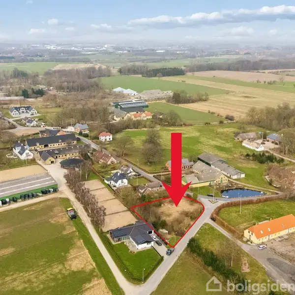 Rødt pile markerer en tom grund omgivet af huse og landbrugsbygninger i et landskab med marker og skov. Grunden ligger tæt på en vej i en landsby.