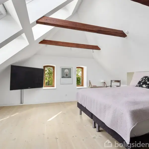 Soveværelse med en stor seng dækket af tæpper, stående i et rum med skråt loft og træbjælker. Fjernsynet er monteret på væggen, ved siden af to små vinduer med udsigt.