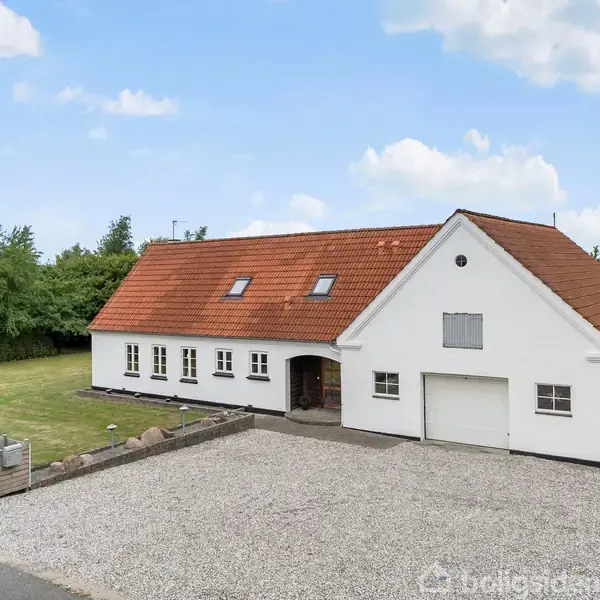 Hvidt hus med rødt tegltag står stille på en grusbelagt indkørsel omgivet af grønne træer og en græsplæne. Der er en garage med en lukket port.