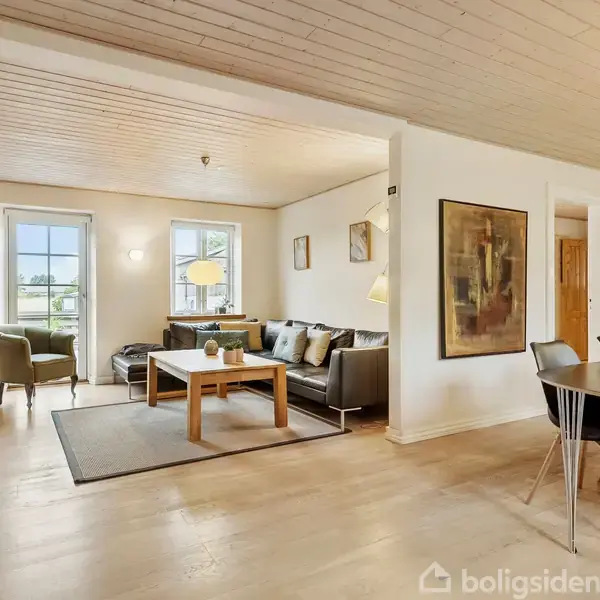 Stue med sofagruppe og TV på væggen, spisestuebord med stole i forgrunden. Rummet er lyst med store vinduer og moderne møbler. Malerier hænger på væggene.