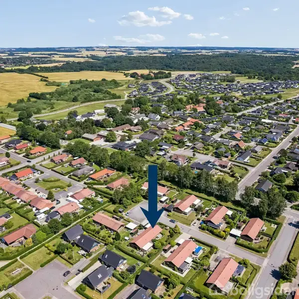 Pil peger på et hus i en forstadsbebyggelse. Husene ligger tæt med velplejede haver. Omgivelserne viser åbne marker og skov i det fjerne under en klar blå himmel.