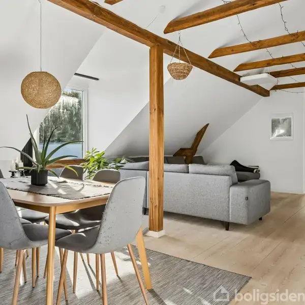 En moderne stue med spisebord og seks stole, placeret nær en skråvæg med vindue. En grå sofa står i baggrunden. Træbjælker pryder loftet, og der er planter på bordet.