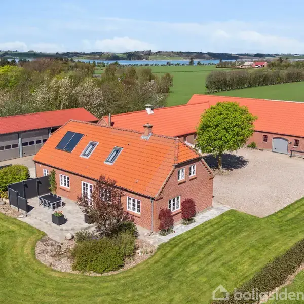 Et rødt murstenshus med orange tag står mellem velplejede græsplæner og træer. Huset har solpaneler og en terrasse foran. Der er også et stort grusareal og en flagstang med dannebrog.