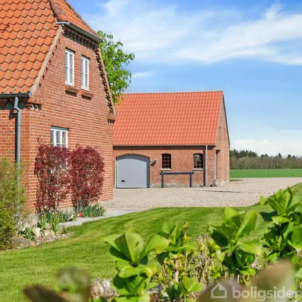 Rødt murstenshus med røde teglstenstag ligger i en frodig have med velplejet græs og buske. I baggrunden ses en garagebygning og en flagstang med et Dannebrog.