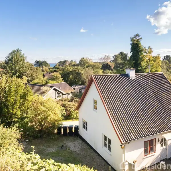 Hus med rødt tag står i en frodig have omgivet af træer og buske. Baggrunden viser flere huse samt en skov og en delvist synlig himmel med spredte skyer.