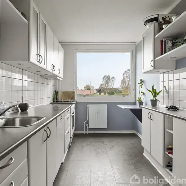 Et moderne køkken med hvide skabe og sorte bordplader. Der er en vask til venstre og en stor vindue i midten, der lukker lys ind fra en udendørs udsigt. Planter og køkkenredskaber dekorerer rummet.