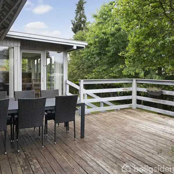 Træterrasse med sort spisebord og seks stole. Bordet er placeret tæt på husets glasindgang. Omkranset af grønne buske og træer, med trægulv og hvidt rækværk.