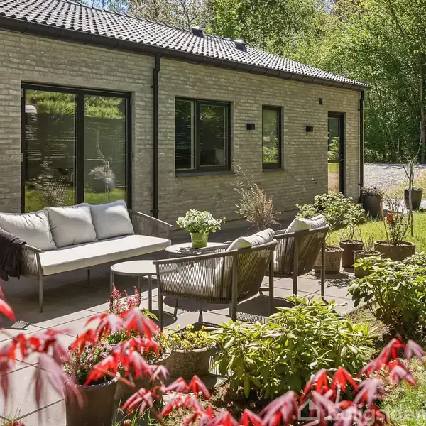 En terrasse med sofa og stole er arrangeret. Omgivet af blomster og planter ved siden af et moderne hus med murstensfacade. I baggrunden ses en frodig have med træer.