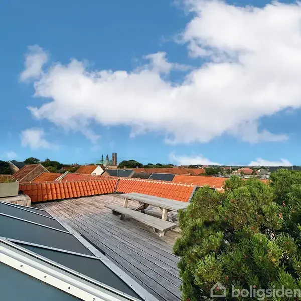 Bord og bænkesæt på en tagterrasse med udsigt over et bylandskab. Røde tegltage og træer omgiver området, og himlen er klar med få skyer.