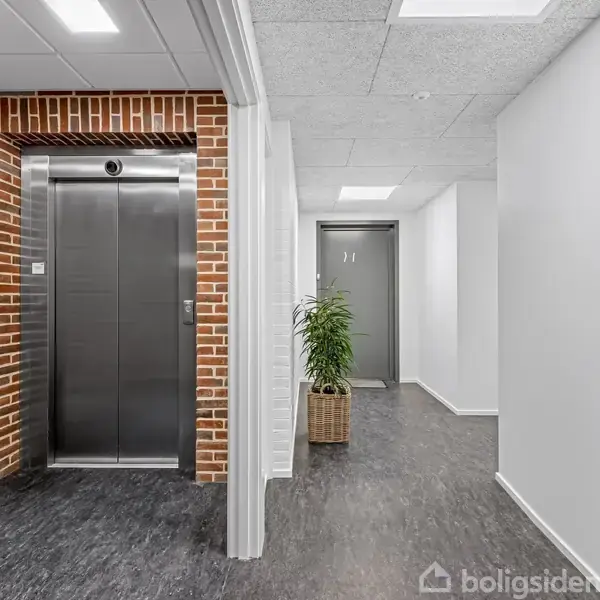 Elevator i ståldør, stationær, ved siden af rødt murværk og indendørs plante; placeret i en lys gang med gråt gulv og hvidmalede vægge.