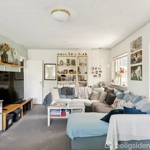 En stor, L-formet grå sofa står i en lys stue med dekorerede vægge og hylder. Et skrivebord med stol er i modsatte ende, og mange puder er på sofaen.