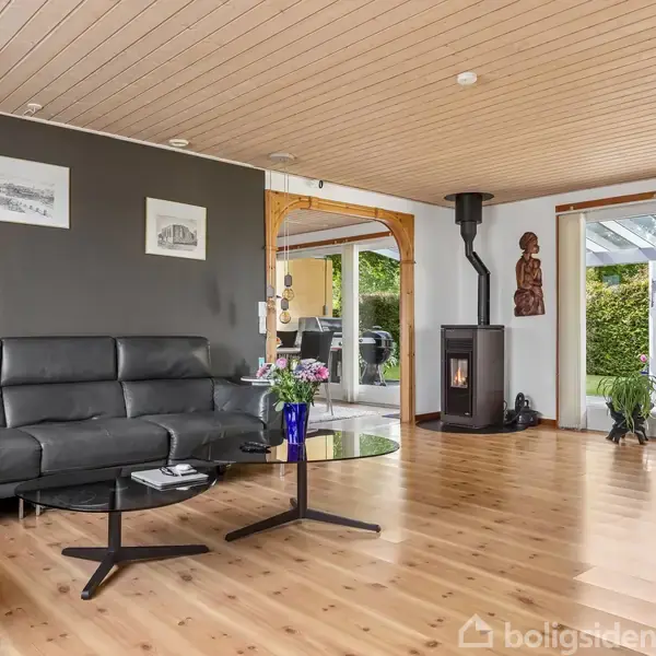 Sort sofa står i en stue med trægulv, grå væg med kunstbilleder. En brændeovn og skulpturer ses ved store vinduer med udsigt til grønne planter.
