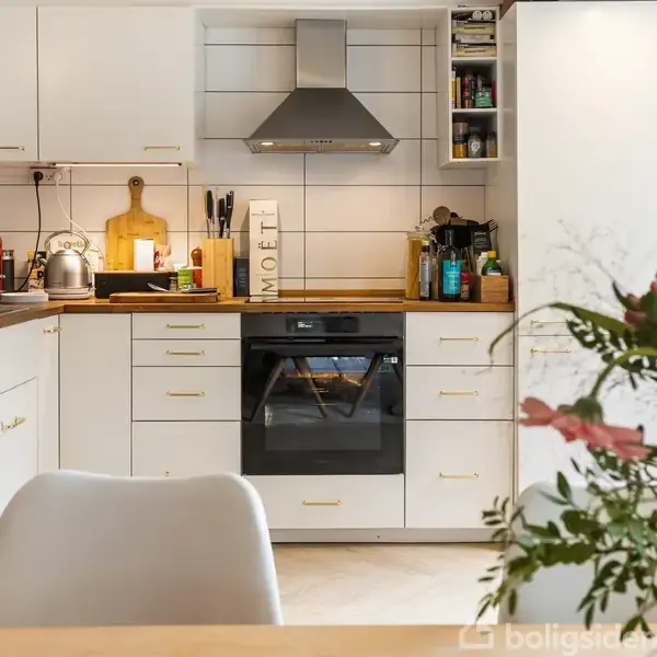 Et køkken med hvide skabe og integreret ovn i midten. En emhætte hænger over ovnen. Blomster står på bordet foran, skaber en hyggelig atmosfære. Tekst: "MOET".