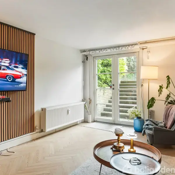 Et moderne stueområde med en vægmonteret fladskærms-tv, der viser en rød sportsvogn i bevægelse. En glasdør fører ud til en terrasse med trapper og grønne planter.