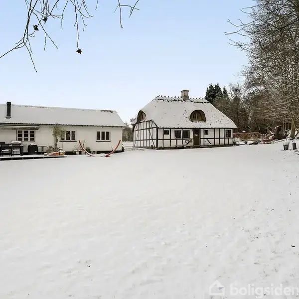 To huse står i et snedækket landskab. Det ene er moderne, mens det andet er bindingsværk. I baggrunden er træer dækket af sne, hvilket skaber en vinterlig atmosfære.