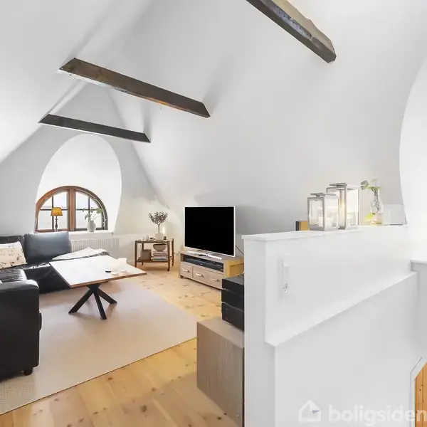 En stue med sort hjørnesofa og sofabord, står opstillet foran et fladskærms-tv. Rummet har skråvægge med synlige bjælker og buede vinduer. Ingen tekst er synlig.