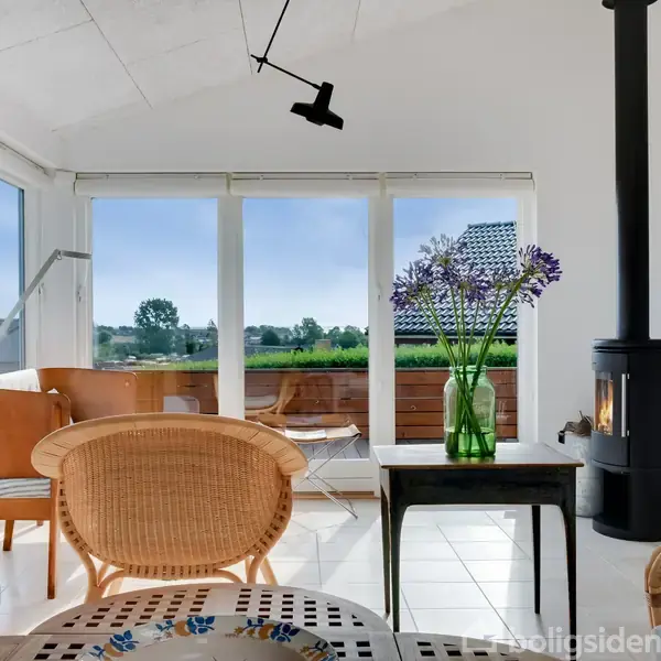 En stue med pejs i gang, bord med vase og blomster, kurvemøbler. Gennem store vinduer ses grønne marker og træer udenfor. Hylder med bøger og dekorative genstande.