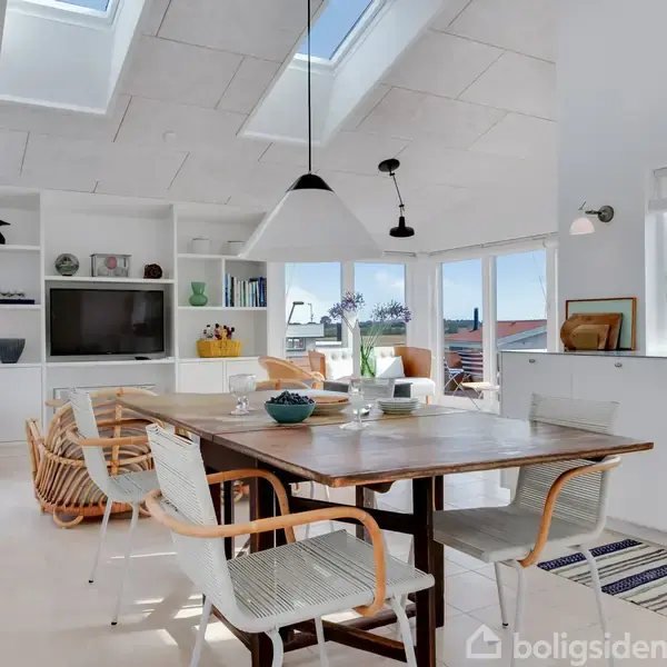 Et moderne spiserum med et træbord omgivet af seks stole. Ovenlysvinduer og store sidevinduer lader sollys strømme ind. Baggrund indeholder en reol med en fladskærm og dekorationer.